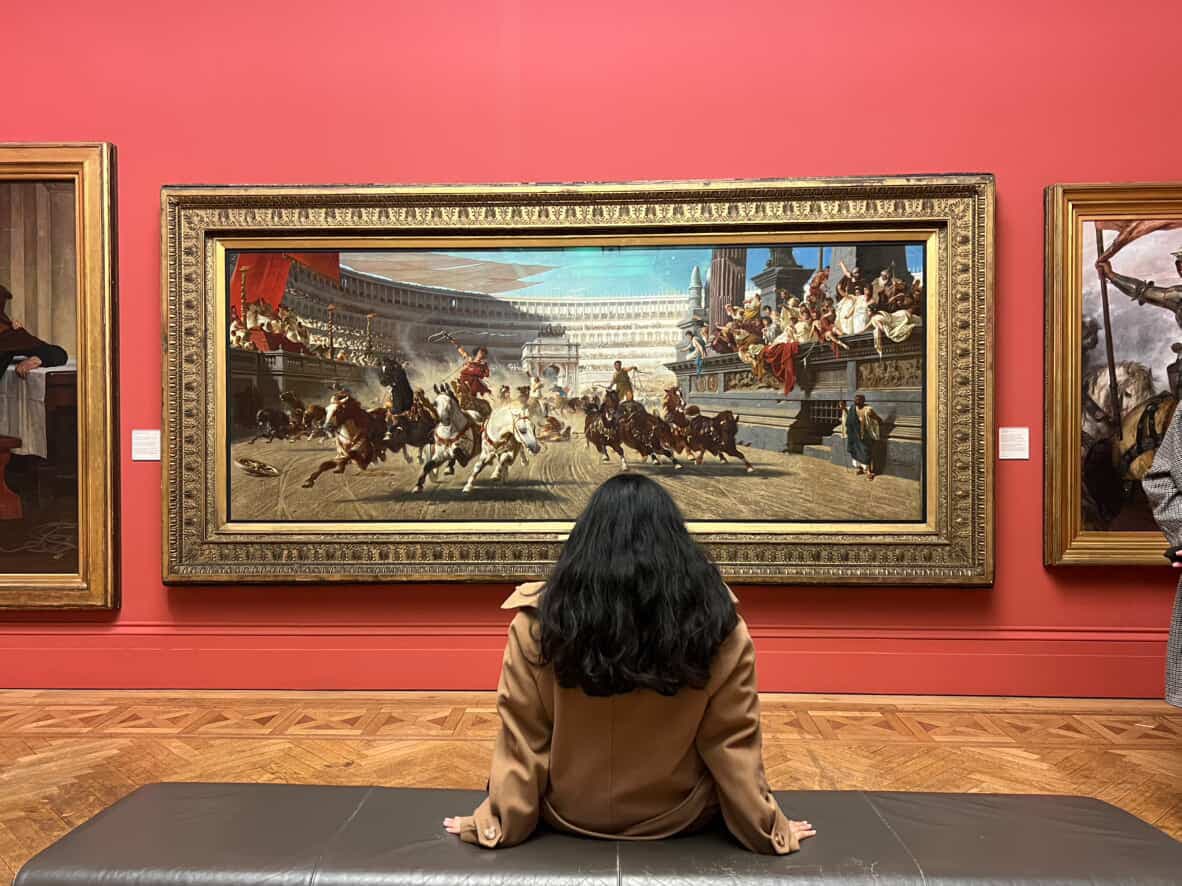 Ananya seated in a gallery, looking at a large painting on a red wall